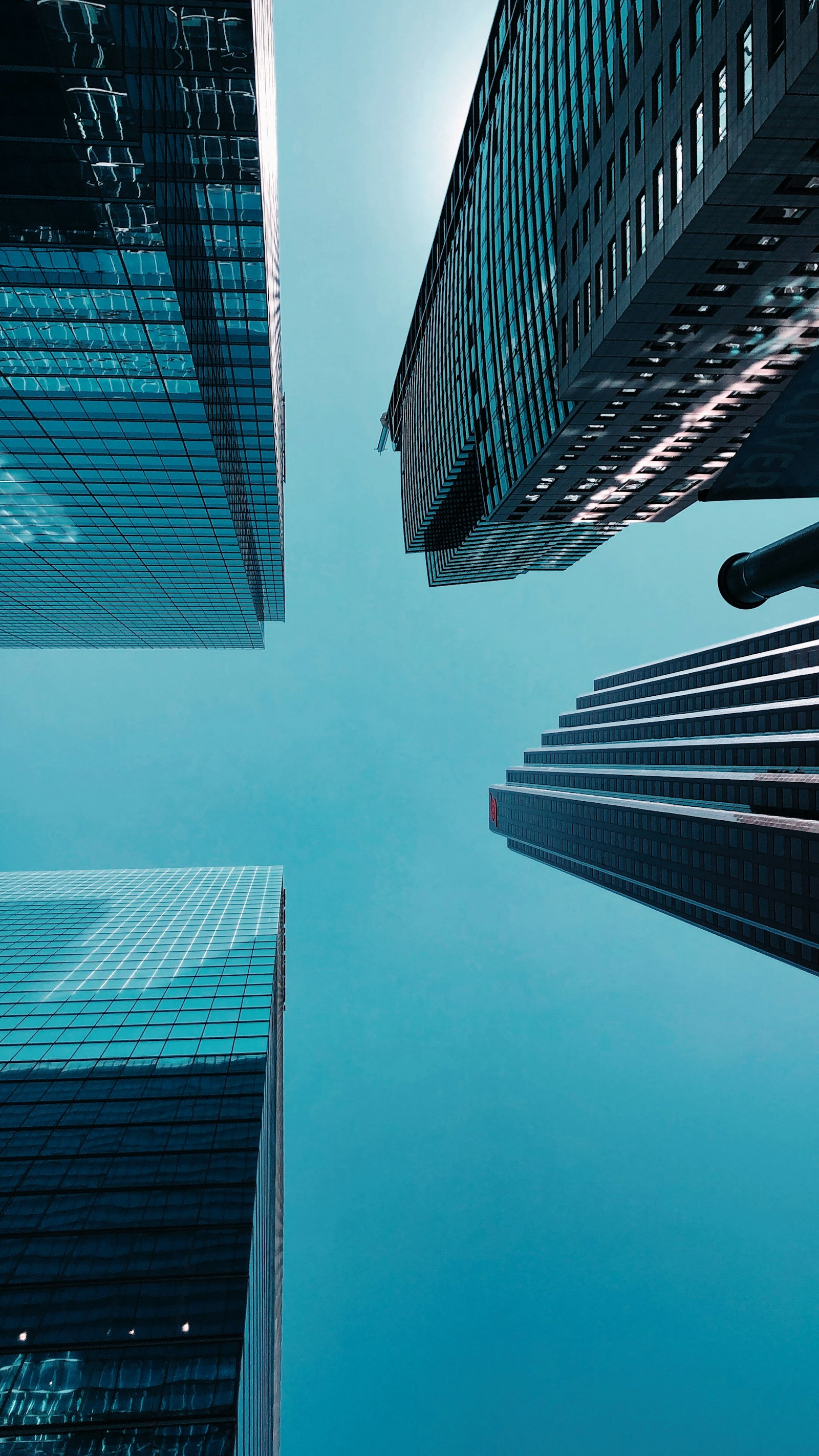 Looking straight up at glass office towers converging against a clear teal sky