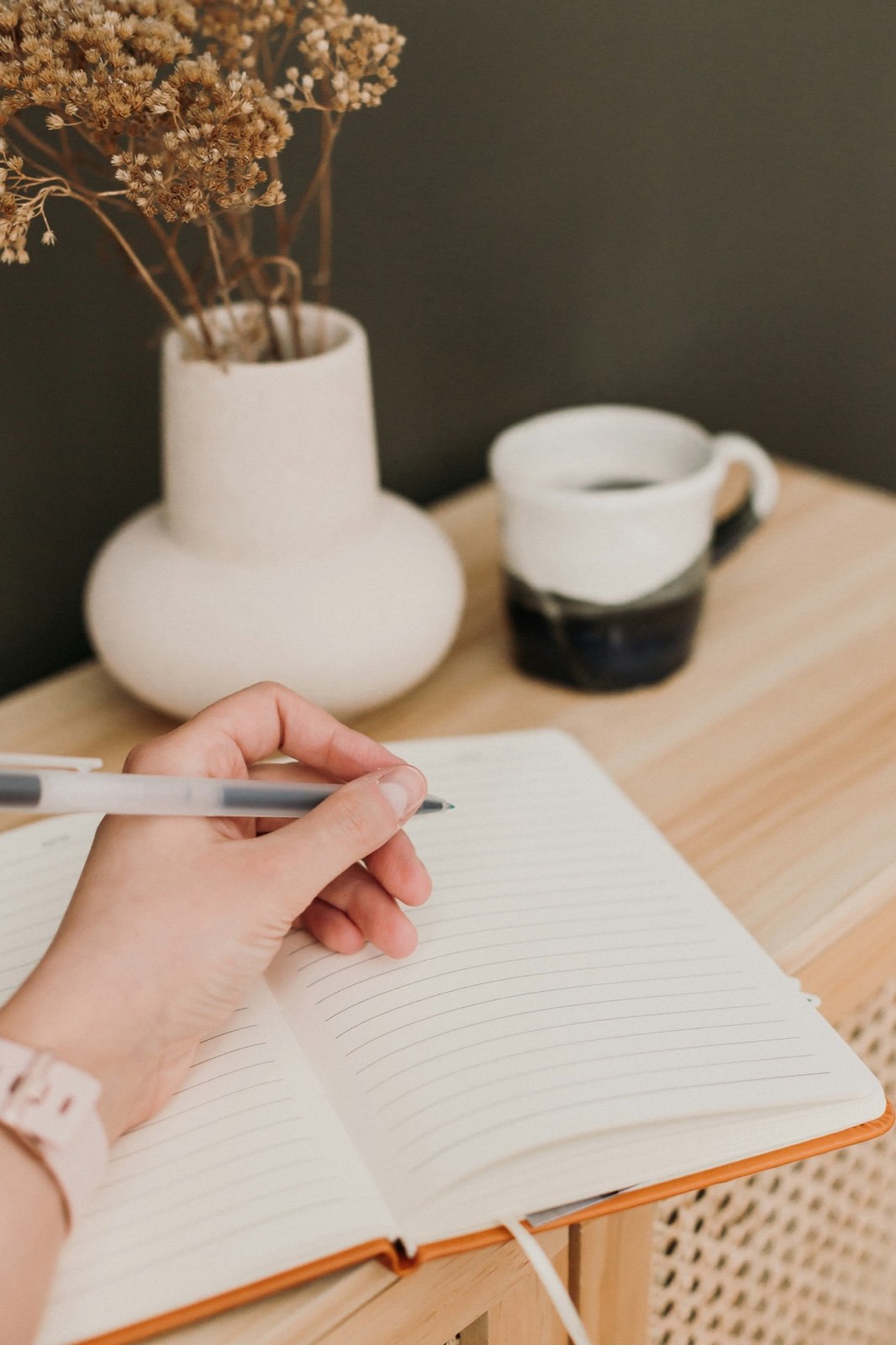 A hand writing in an open notebook next to a cup of coffee and a vase of dried flowers on a wooden desk